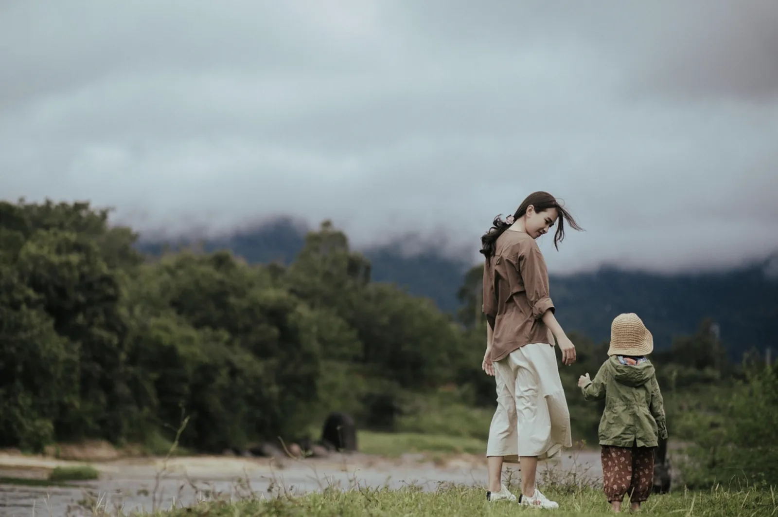 A mother and young child standing beside a river with green hills and low clouds in the background