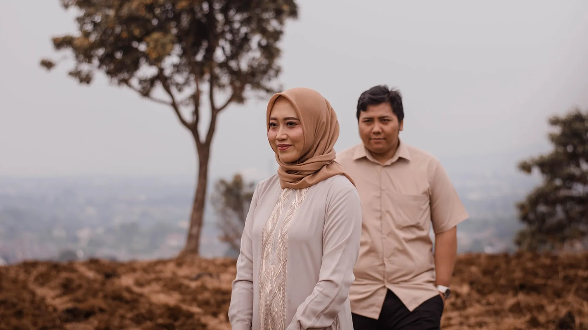A South Asian couple standing outdoors against a hazy backdrop of distant city and hills