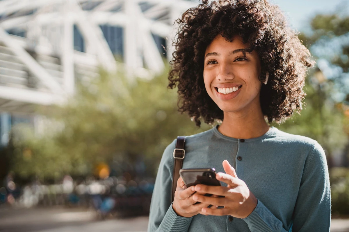 A smiling woman holding her phone on a sunny city street