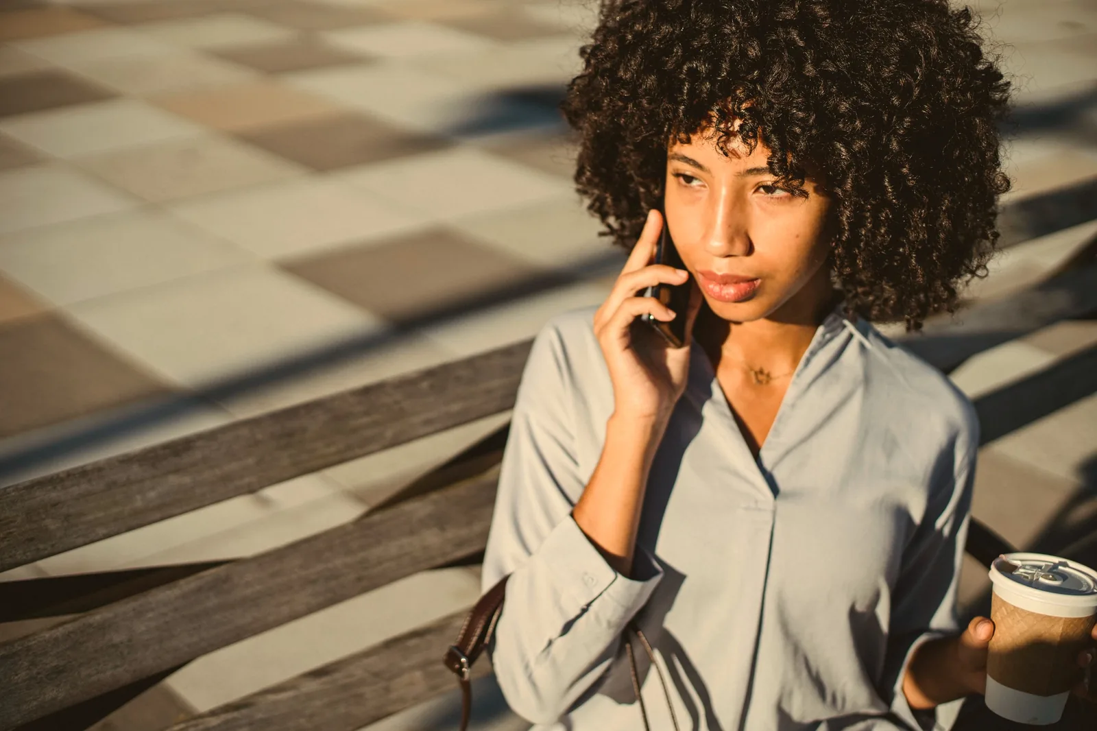 A woman with curly hair talking on her phone in afternoon sun, holding a coffee